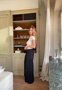 Haut blanc à manches courtes et pantalon noir ample à taille haute. Étagères en bois minimalistes avec des objets décoratifs en arrière-plan.