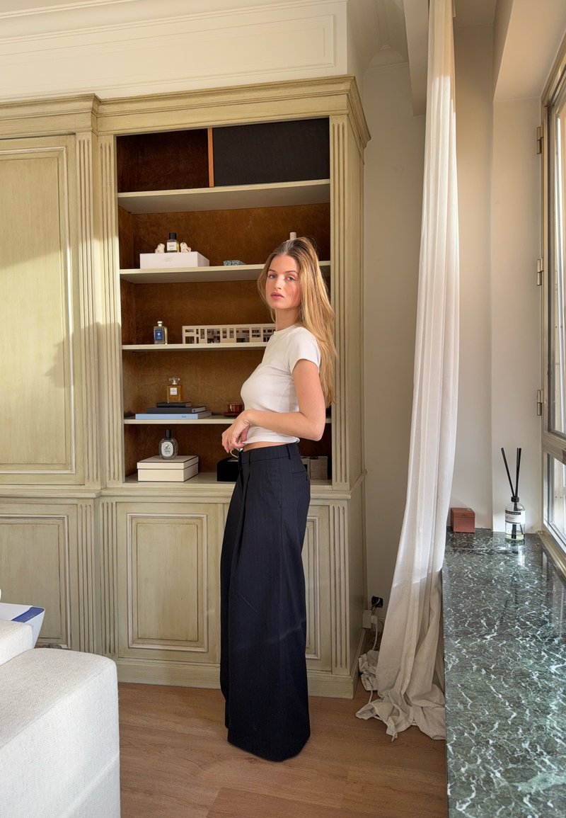 Haut blanc à manches courtes et pantalon noir taille haute à jambes larges. Étagère en bois minimaliste avec des objets décoratifs en arrière-plan.