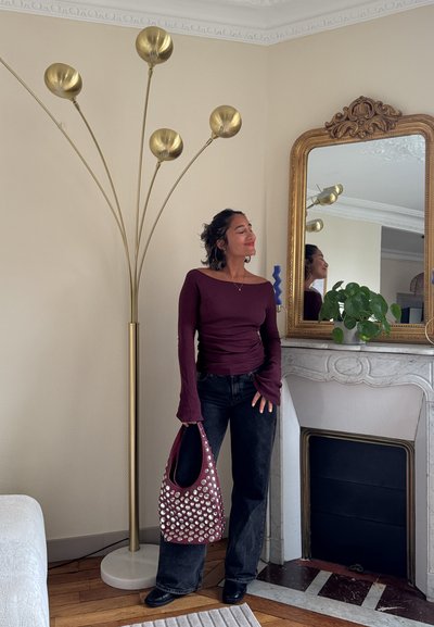 Mujer con blusa burdeos y jeans negros, sosteniendo un bolso con tachuelas, de pie junto a una lámpara de pie alta dorada y un espejo ornamentado sobre una chimenea blanca con una planta.