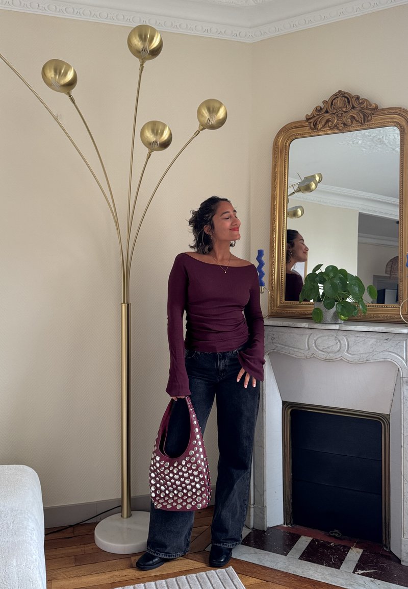Woman in burgundy top and black jeans holding studded bag, standing near tall gold floor lamp and ornate mirror above white fireplace with plant.