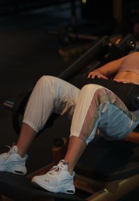 Person using leg press machine, wearing white sneakers and white track pants with colored stripes in a dimly lit gym.