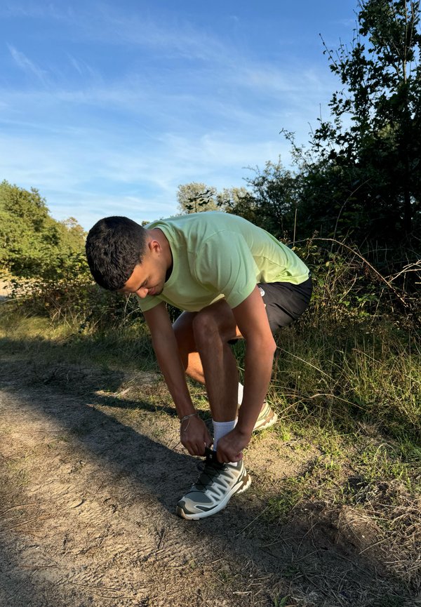 Una persona è in ginocchio su un sentiero di terra, mentre allaccia delle scarpe da esterno grigie e bianche con dettagli verdi. Indossa una maglietta verde chiaro e pantaloni corti neri.