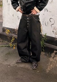 Personne portant un jean noir oversize à fermeture à boutons, des chaussures en cuir marron et une veste noire avec des poignets en polaire, debout près d'un mur de graffiti.