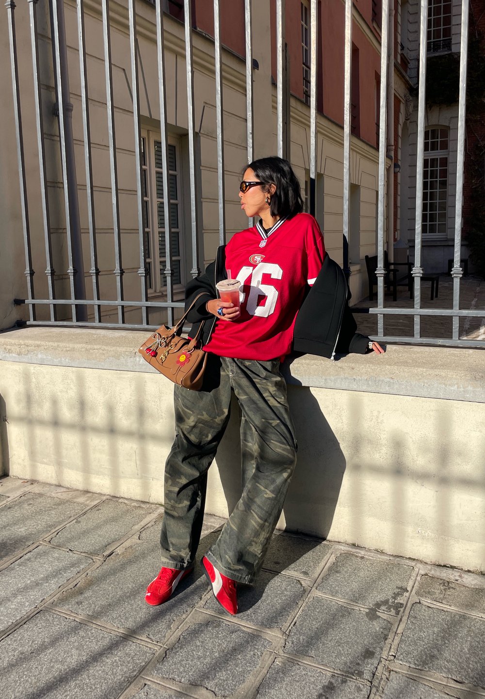 Rød fotballtrøye med hvitt nummer, svart oversized jakke, camo-bukser og røde joggesko. Brun håndveske med dekorative detaljer.