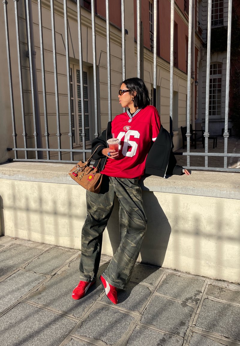 Červený futbalový dres s bielym číslom, čierna oversized bunda, maskáčové nohavice a červené tenisky. Hnedá kabelka s dekoratívnymi prvkami.