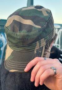 Camouflage-patterned cap in green and brown fabric with a curved brim; features stitching details and a minimalist logo on the back.