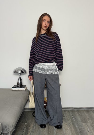 Mujer de pie en el interior, vistiendo una camisa de mangas largas a rayas y pantalones anchos grises con detalle de encaje blanco, sosteniendo un bolso beige junto a un sofá y una lámpara.