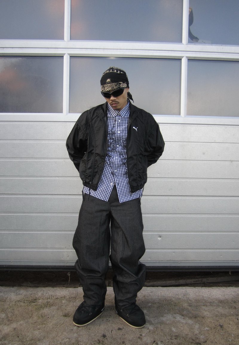 Giacca nera leggera sopra una camicia a quadri blu e bianca, con pantaloni grigi larghi e scarpe da ginnastica nere, in piedi davanti a una porta del garage.