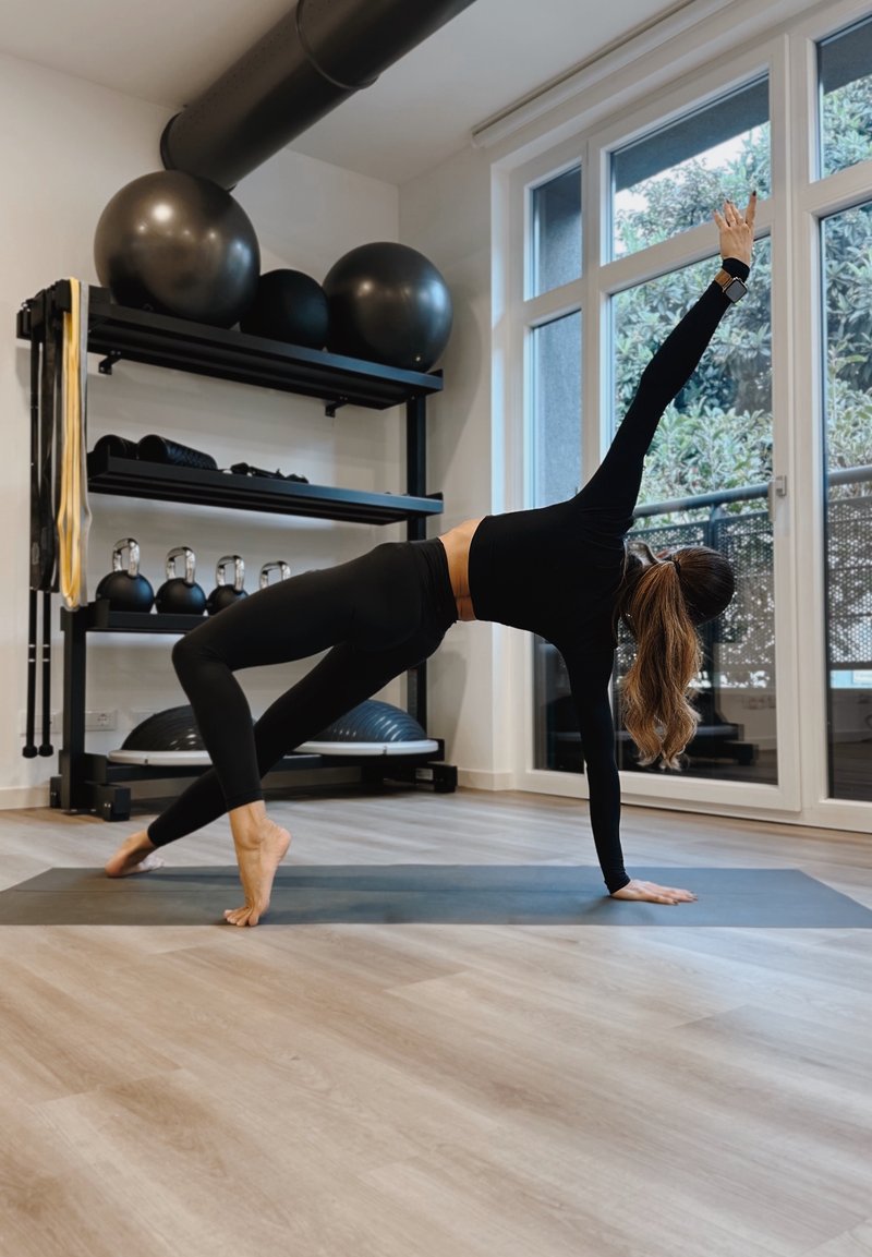 Žena ve černém sportovním oblečení balancuje na jedné ruce a jedné noze na jógové podložce v jasné tělocvičně s cvičebními míči a kettlebelly.