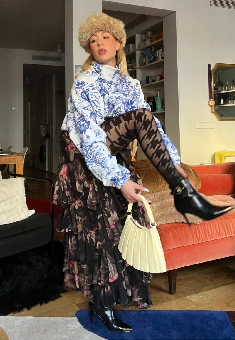 Femme à l'intérieur portant une jupe fleurie, une veste bleue et blanche à motifs, un chapeau en fourrure, des collants à motifs, des bottes à talons noires, tenant un sac à main crème.