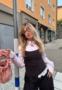 Noor blonde juustega noor naine kannab kihilist roosa pluusi ja musta toppi, hoides roosat kotti, seistes linnatänaval posti lähedal.