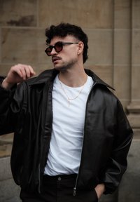 Schwarze Jacke aus Kunstleder mit weitem Kragen, darunter weißes T-Shirt, goldene Kettenhalskette und runde schwarze Sonnenbrille. Dunkle Hose.