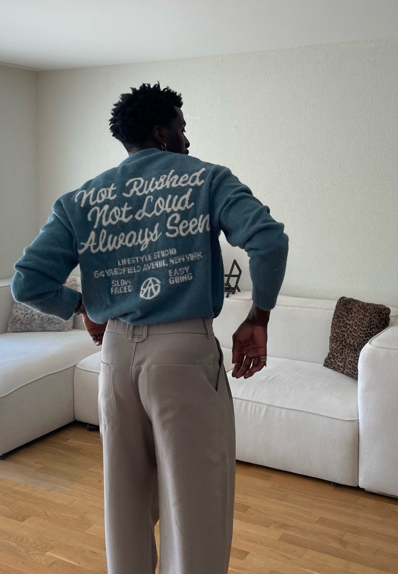 Homme en pantalon beige et pull bleu avec texte blanc debout dans un salon avec canapé blanc et coussin imprimé léopard.