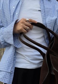 Osoba v bielom tričku a modrej kockovanej košeli drží hnedé semišové držadlá tašky s dvomi striebornými prsteňmi na ľavej ruke.