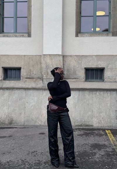 Joven con cuello alto negro y pantalones, de pie en una calle frente a un muro de piedra con ventanas, cubriéndose parte del rostro con una mano.