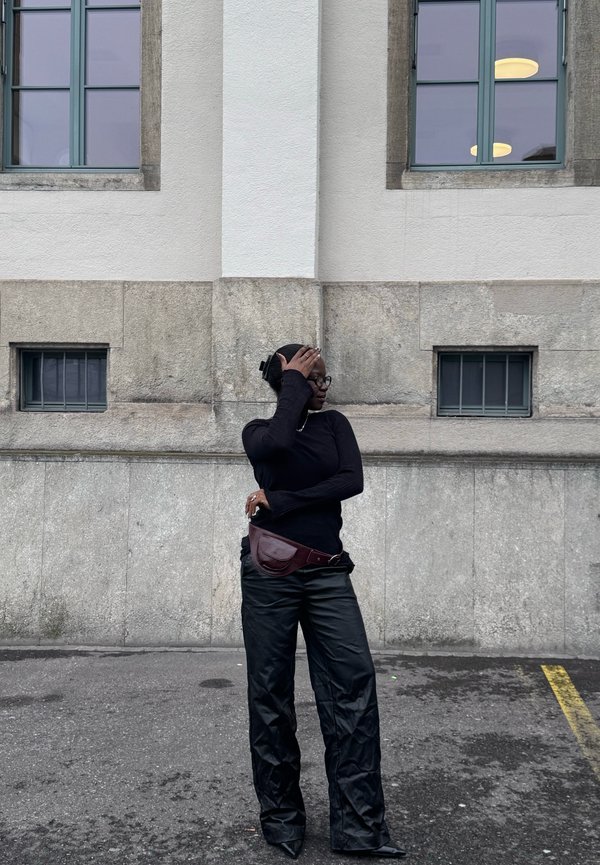 Tânăr îmbrăcat într-un helancă neagră și pantaloni, stând pe o stradă în fața unui zid de piatră cu ferestre, acoperindu-și o parte a feței cu o mână.