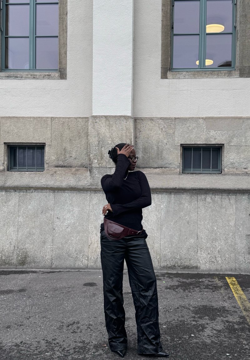 Jeune personne portant un col roulé noir et un pantalon, debout dans une rue devant un mur en pierre avec des fenêtres, couvrant une partie de son visage avec une main.