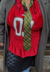 Červená saténová košeľa s nariaseným pásom, s veľkým bielym číslom, kombinovaná so zelenou károvanou kravatou. Nosená pod sivým blejzrom a čiernou čipkovanou sukňou.