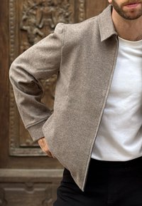 Braune Jacke aus Wollmischgewebe mit Kragen und Reißverschluss, kombiniert mit einem weißen Hemd, vor einem strukturierten Holzhintergrund.