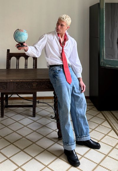 Camisa blanca de manga larga, corbata roja, vaqueros azules holgados y botas negras hasta el tobillo. Sosteniendo un pequeño globo. Mesa de madera oscura y suelo con un patrón.