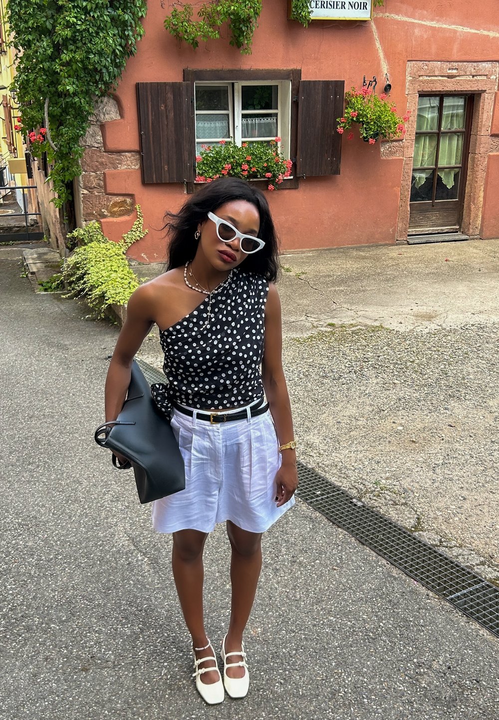 Μαύρο και λευκό τοπ με πουά σχέδιο στον έναν ώμο, συνδυασμένο με λευκές λινές βερμούδες, μαύρη τσάντα και λευκά παπούτσια με φάσες στον αστράγαλο.