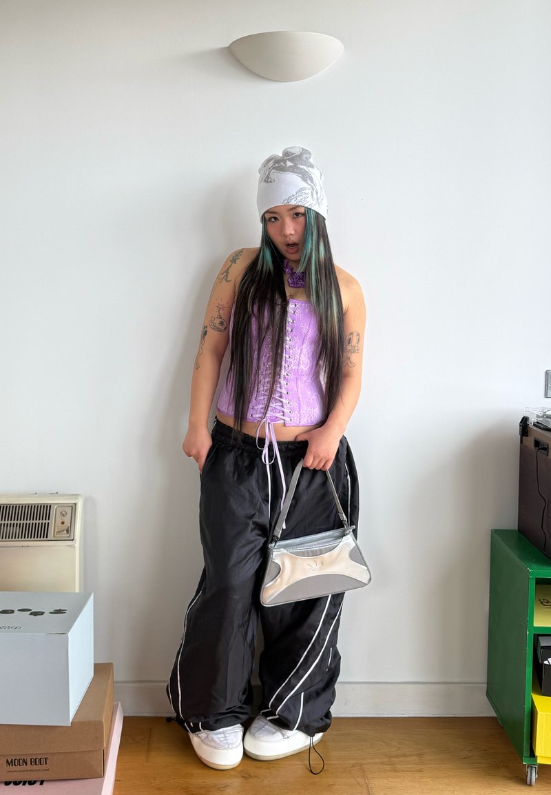 Jeune personne aux longs cheveux portant un bonnet blanc, un corset violet, un pantalon noir ample, tenant une pochette argentée, debout à l'intérieur contre un mur blanc.