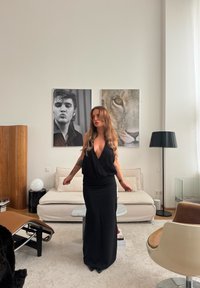 Zwarte jurk met een diepe V-hals, aansluitend lijfje en vloeiende rok. Licht houten meubels, een witte sofa en kunst aan de muren vullen de ruimte aan.