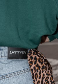 Zelená mikina, světle modré džíny, černý opasek s logem "URTRN" a hnědý šátek s tygřím vzorem uvázaný v pase.