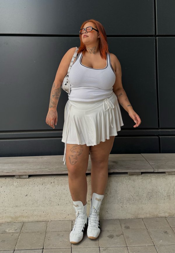 Žena s červenými vlasmi, ktorá má na sebe slnečné okuliare, biele tielko, sukňu a biele vysoké tenisky, stojaca pri tmavom stenovom obklade.