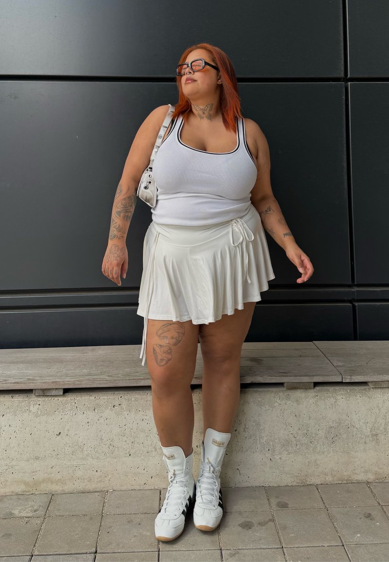 Frau mit roten Haaren, die Sonnenbrille, ein weißes Tanktop, einen Rock und weiße High-Top-Sneaker trägt, steht an einer dunklen Wand mit Paneelen.