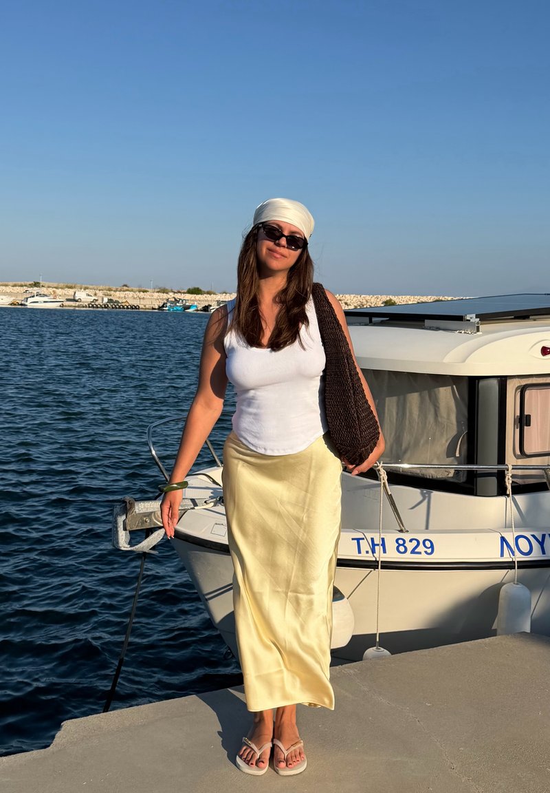 Γυναίκα με γυαλιά ηλίου, λευκό τοπ, κρεμ φούστα και μαντήλι στο κεφάλι στέκεται σε προβλήτα δίπλα σε αγκυροβολημένο σκάφος κάτω από δυνατό ηλιακό φως κοντά σε ήρεμα νερά.