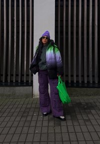 Φουσκωτό μπουφάν με κλίση από πράσινο σε μωβ, συνδυασμένο με σκούρο πράσινο φούτερ, μωβ παντελόνι cargo και φωτεινή πράσινη τσάντα. Μαύρα παπούτσια.