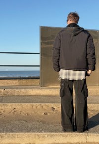 Doudoune noire avec une capuche texturée, portée sur une chemise grise à carreaux effilochée, associée à un pantalon cargo noir ample avec des poches.