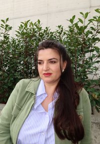 Vrouw met lang donker haar en rode lippenstift, gekleed in een groene jas en gestreept shirt, staande voor groene, bladrijk struiken.