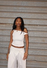 Witte fitted top met een off-the-shoulder ontwerp en bijpassende hoge taille broek met plooien. Gouden sieraden accenten. Grijze achtergrond.