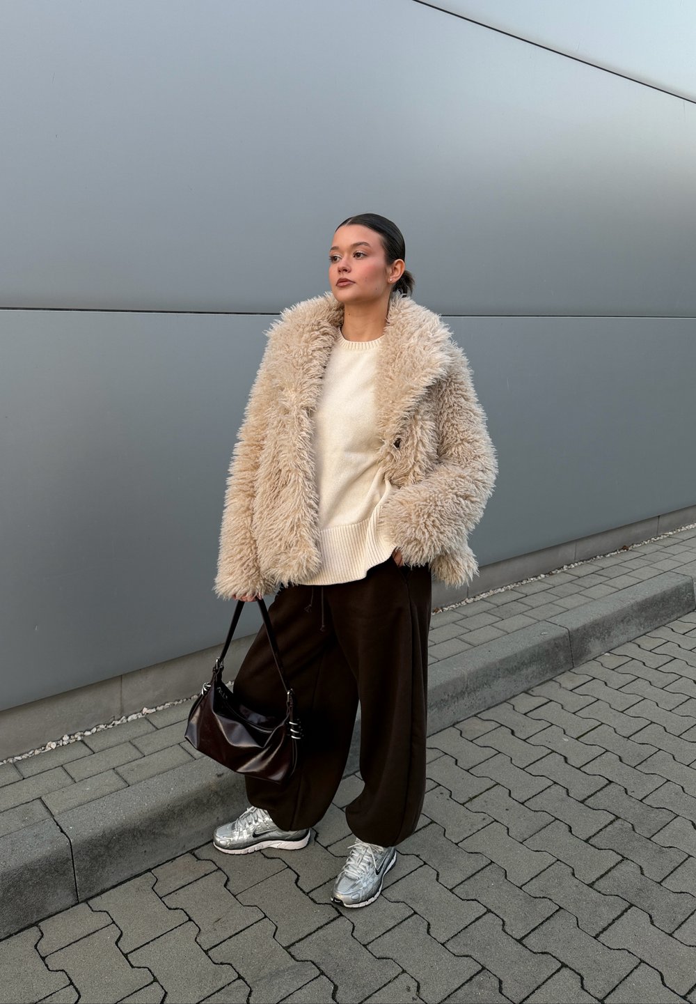 Beige faux fur jacket over a cream jumper, paired with loose brown trousers and silver trainers. A black handbag completes the look.