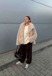 Beige faux fur jacket over a cream jumper, paired with loose brown trousers and silver trainers. A black handbag completes the look.