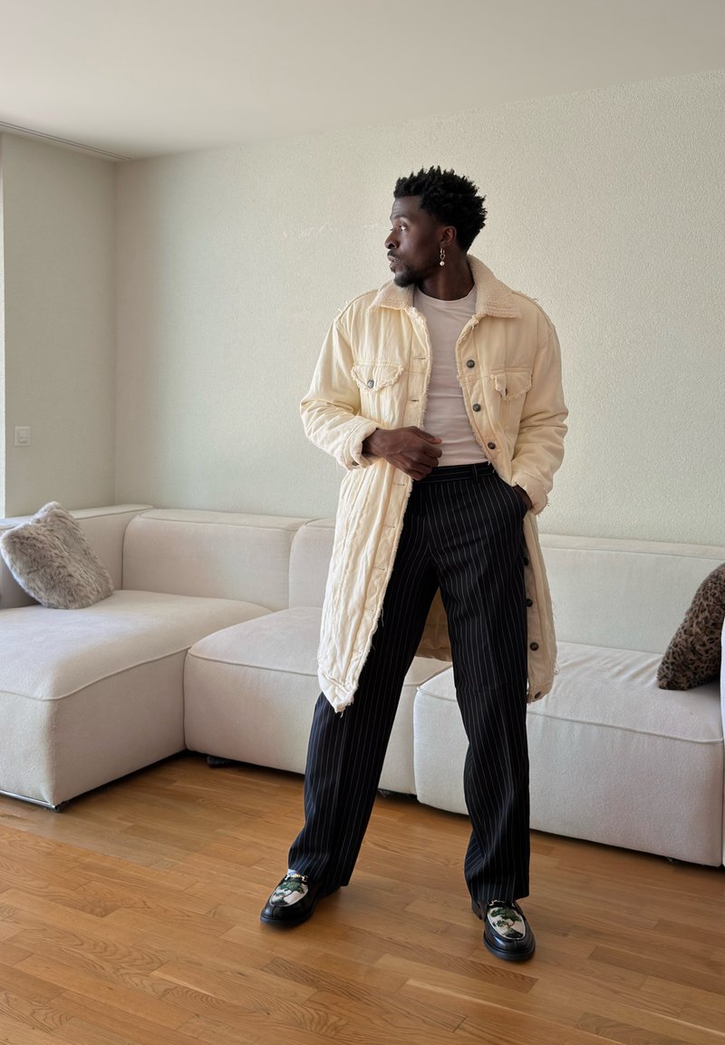 Homme portant une longue veste couleur crème, une chemise blanche, un pantalon à rayures fines et des chaussures à motifs, debout dans un salon à côté d'un canapé beige.