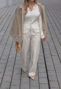 Bēša vilnas wraps ar bārkstīm, krēmkrāsas blūze ar pogām, krēmkrāsas plašas bikses un šauras bēša krāsas augstpapēžu kurpes. Rokās tur brūnas aušanas somu.