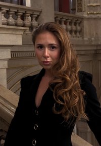 Czarna, dopasowana marynarka z głębokim dekoltem w kształcie litery V i błyszczącymi srebrnymi guzikami. Długie, falowane brązowe włosy; miękka skóra i naturalny makijaż; tło z marmurowymi schodami.