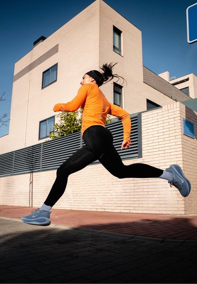Mujer con chaqueta naranja y leggings negros corriendo al aire libre en una zona urbana con edificios modernos de color beige y cielo azul despejado.