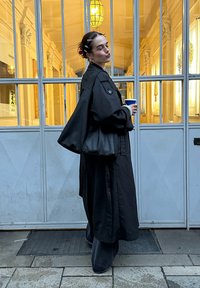 Trench-coat noir ample, superposé à un pantalon à jambes larges. Tissu texturé, fermeture à boutons sur le devant, et manches surdimensionnées. Tenant une tasse.