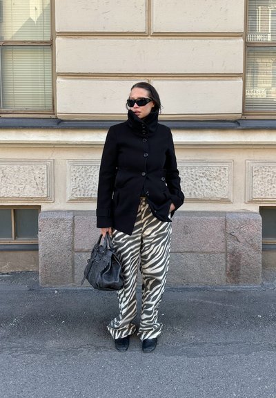 Mujer vestida con abrigo negro, pantalones a rayas de cebra, zapatos negros y gafas de sol, sujetando un bolso negro, de pie junto a una pared de piedra beis.