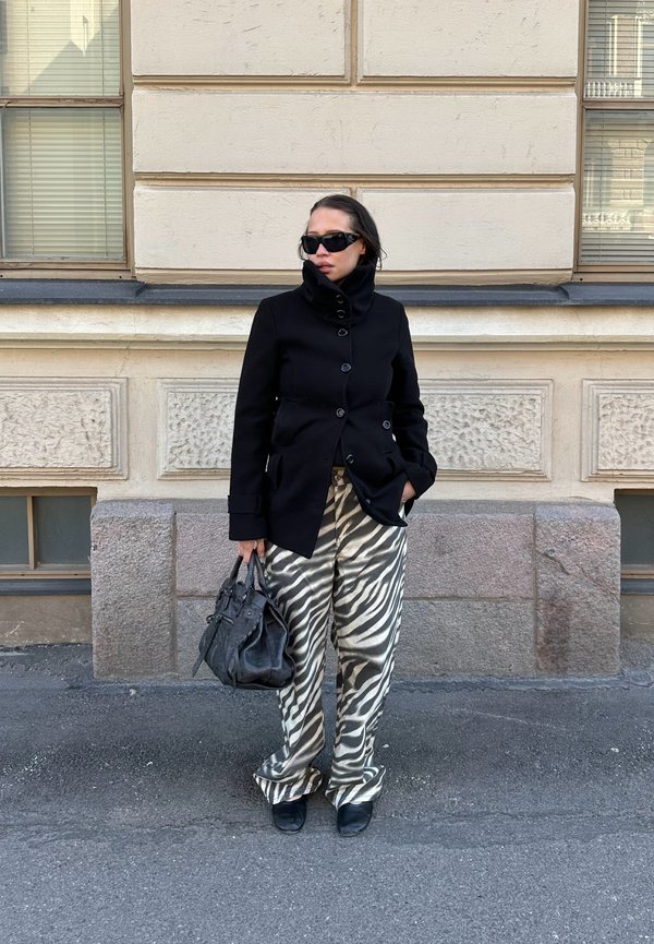 Žena nosi crni kaput, hlače u zebrine pruge, crne cipele i sunčane naočale, drži crnu torbu, stoji uz bež kamen zid.