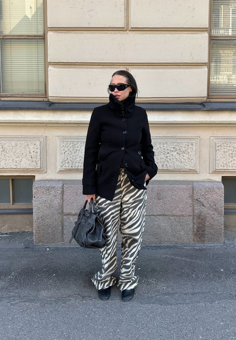 Frau trägt schwarzen Mantel, Zebra-gestreifte Hose, schwarze Schuhe und Sonnenbrille, hält eine schwarze Handtasche und steht an einer beigen Steinmauer.