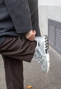 Harmaa lenkkikenkä, jossa verkkokangasta ja synteettisiä materiaaleja, näkyvä musta swoosh-logo, punaiset yksityiskohdat ja kuvioitu pohja, yhdistetty ruskeiden housujen kanssa.