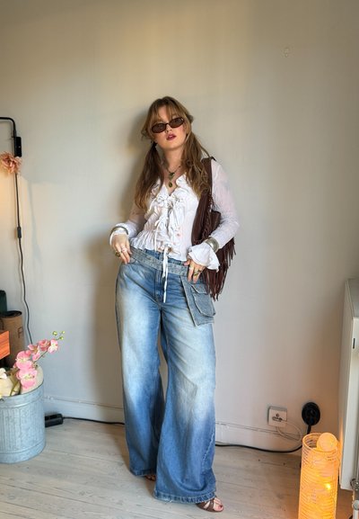 Blusa blanca con volantes y mangas largas, jeans de pierna ancha azul claro, joyería en capas y un bolso marrón con flecos. Iluminación suave y plantas de fondo.