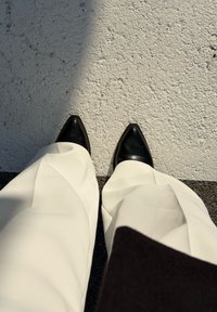 Pantofi cu toc ascuţit negri, combinați cu pantaloni albi cu talie largă, văzuți de sus pe o suprafață texturată, de culoare deschisă.