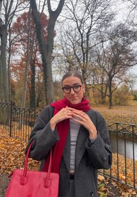 Sivko plašč z večjimi rokavi, rdeča ruta, srebrni prstani in sijoča rdeča torbica. Ozadje prikazuje jesenska drevesa in ribnik.