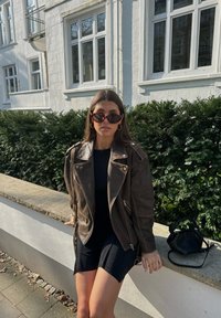 Rjava usnjena motoristična jakna s srebrnimi dodatki, črna prilagojena obleka, črna torbica, temna sončna očala in gladka stenska površina.